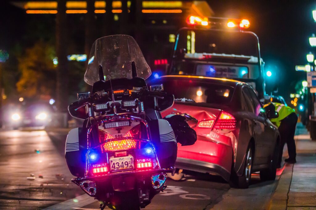 A law enforcement motorcycle is parked behind a car accident scene on the side of a busy road at night.