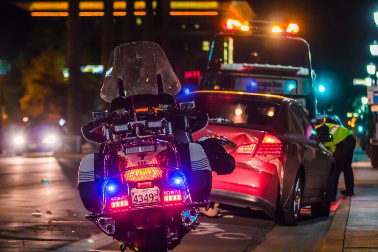 A law enforcement motorcycle is parked behind a car accident scene on the side of a busy road at night.