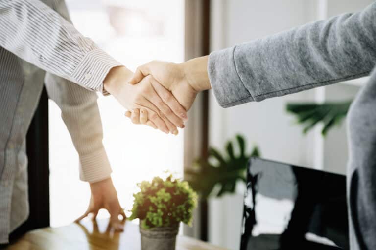 Two people shaking hands in a bright office, with a potted plant on the desk.