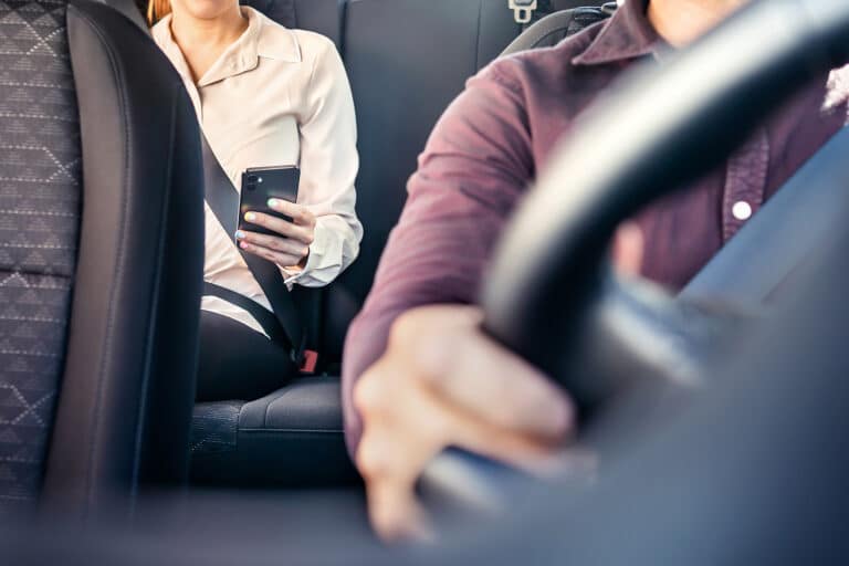 Passenger in back seat wearing seatbelt and using smartphone while driver focuses on the road inside a car.