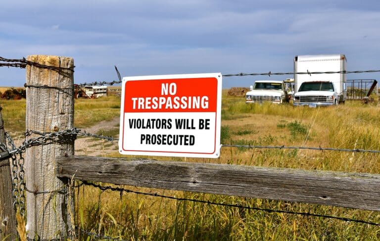 No trespassing sign on a barbed wire fence warning violators, with old trucks and a rural field in the background.