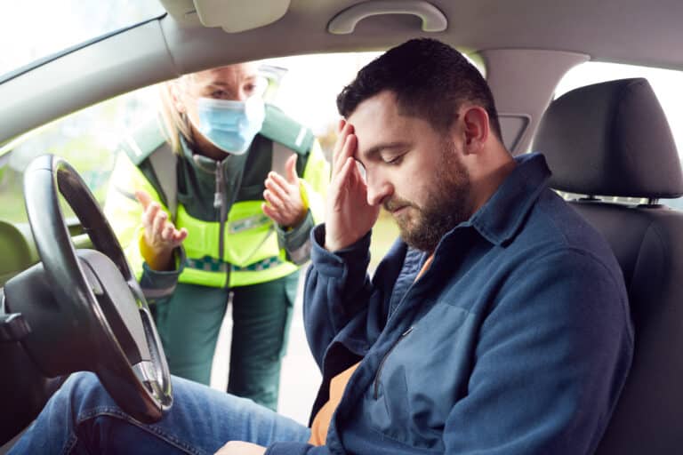 Distressed man in a car holding his head as a female paramedic in a mask talks to him through the window.