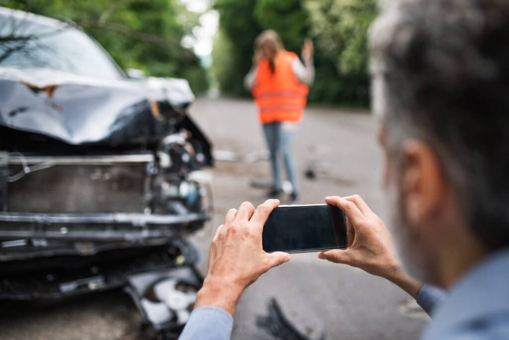 Man taking photo of damaged car after crash, while woman in safety vest gestures in background on empty road.