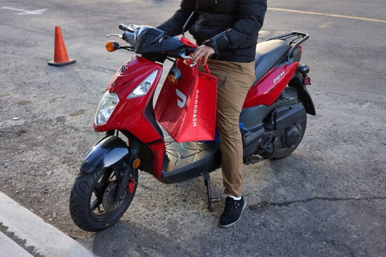 Person on a red scooter holding a DoorDash delivery bag, preparing to make a food delivery in an urban parking area.