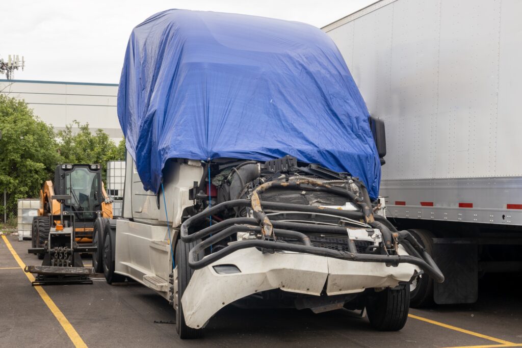 Damaged semi-truck with a blue tarp covering the cab, parked next to a trailer in a lot.