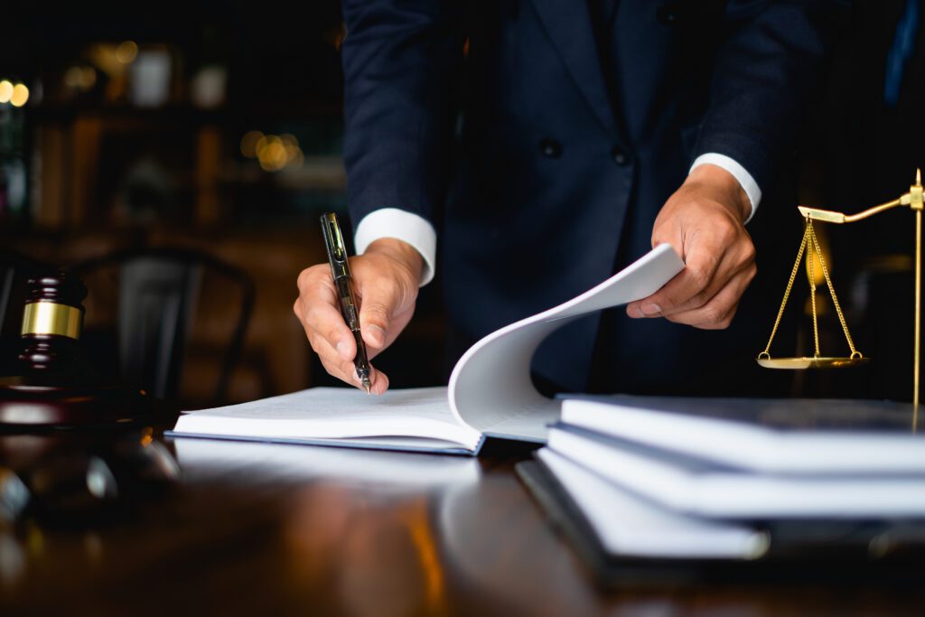 A person in a suit signing documents at a desk with a gavel and scales of justice nearby.