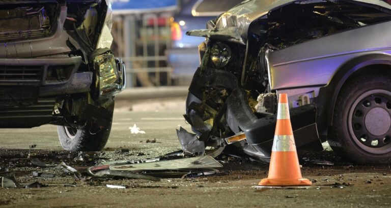 Two damaged cars in a crash with a traffic cone on the ground, debris scattered around.