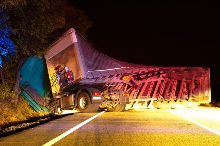 Truck overturned on road at night, blocking lane with significant damage and debris scattered.