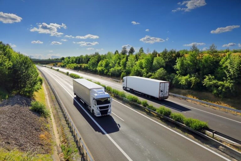 Two trucks driving on a sunny highway surrounded by green trees and blue sky.