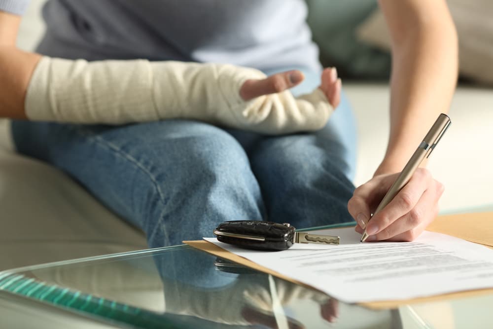 Close-up of woman's hands with broken arm signing insurance papers post-car accident at home.