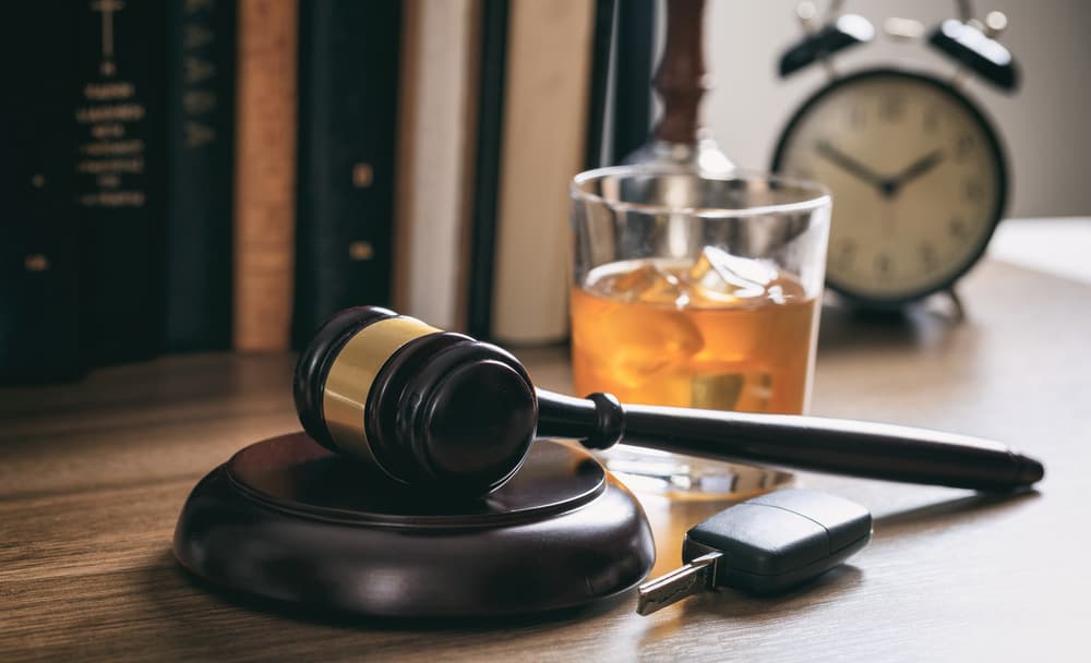  A wooden desk is depicted against a dark background, upon which rests a law gavel, a bottle of alcohol, and a set of car keys. The arrangement symbolizes the dangerous combination of alcohol consumption and driving, highlighting the legal consequences associated with this reckless behavior.