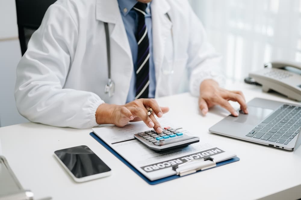 The concept of healthcare costs and fees: A doctor, equipped with a calculator and smartphones or tablets, calculates medical expenses at a hospital. 