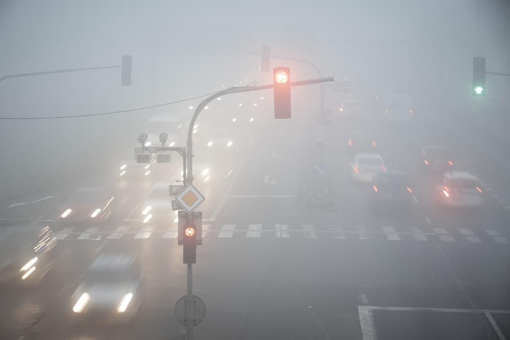 At the road intersection, thick fog blankets the scene, creating blurred motion as cars navigate through the haze.