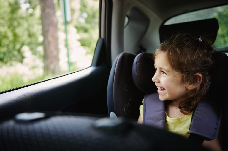 A child sits safely secured in a car seat while looking out the window and smiling as the vehicle moves