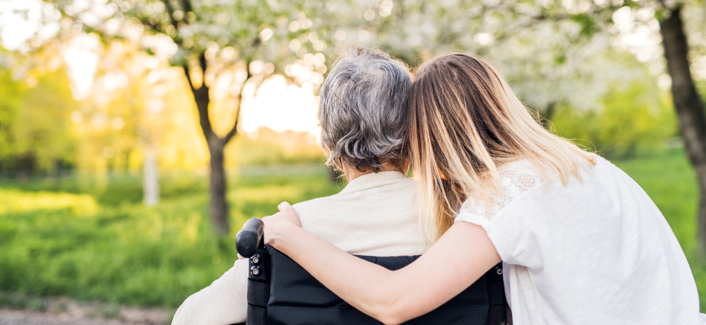 an elderly person sits on a chair with a loved one hugging their shoulders, both people face a natural scene of trees and greenery