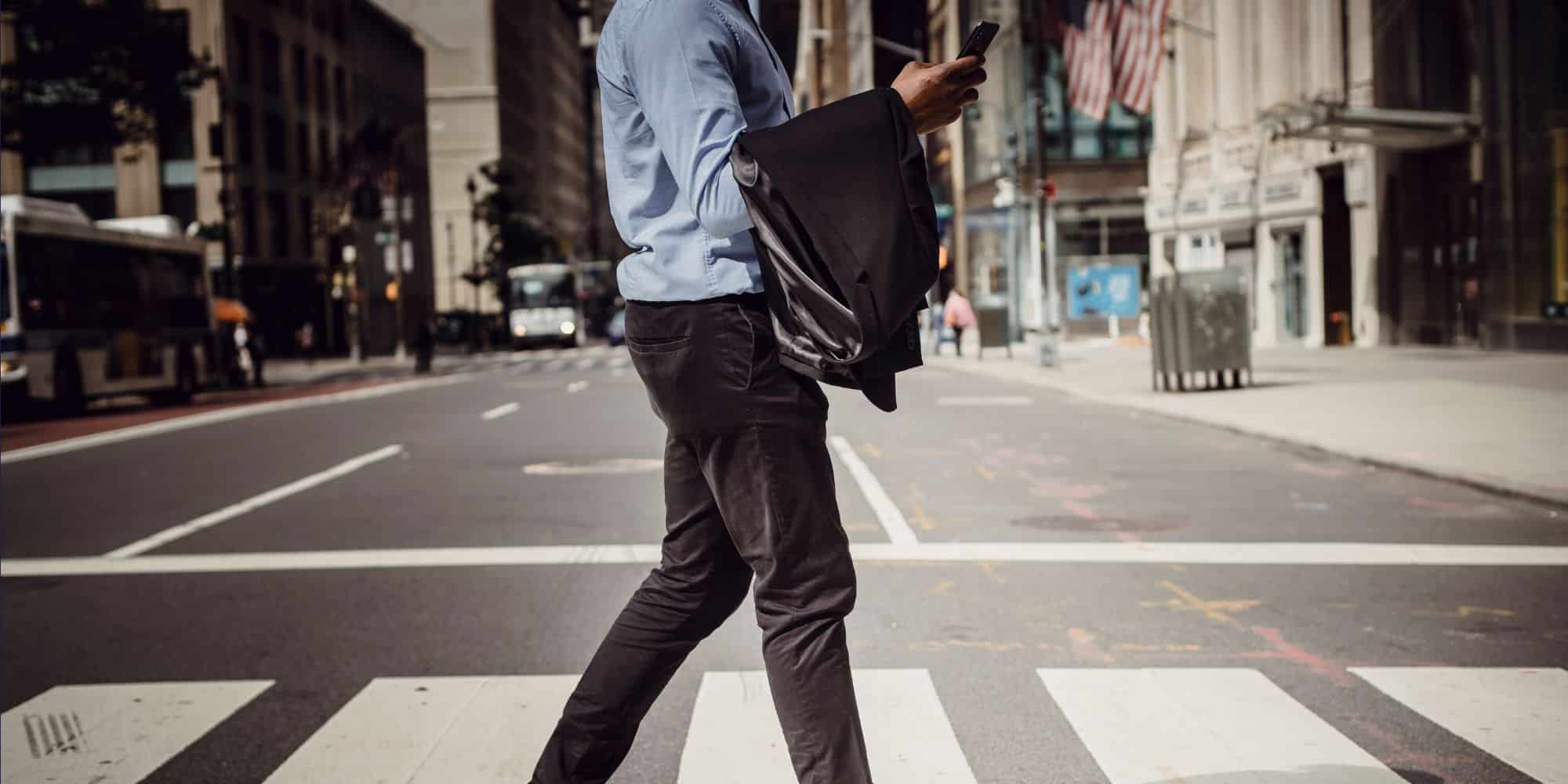 a pedestrian in business casual attire walks on a crosswalk carrying their jacket and phone, there is a cityscape in the background