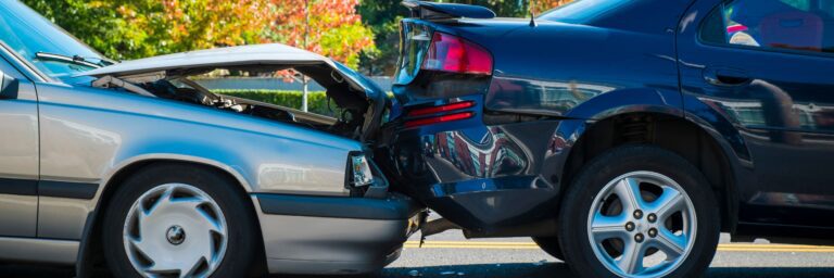 Un automóvil plateado choca por detrás a un automóvil negro, causando un accidente automovilístico. Esto ocurre en medio de una calle en un día soleado.