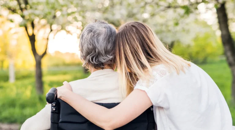 an elderly person sits in a chair looking at greenery, their loved one sits next to them with an arm around their shoulders