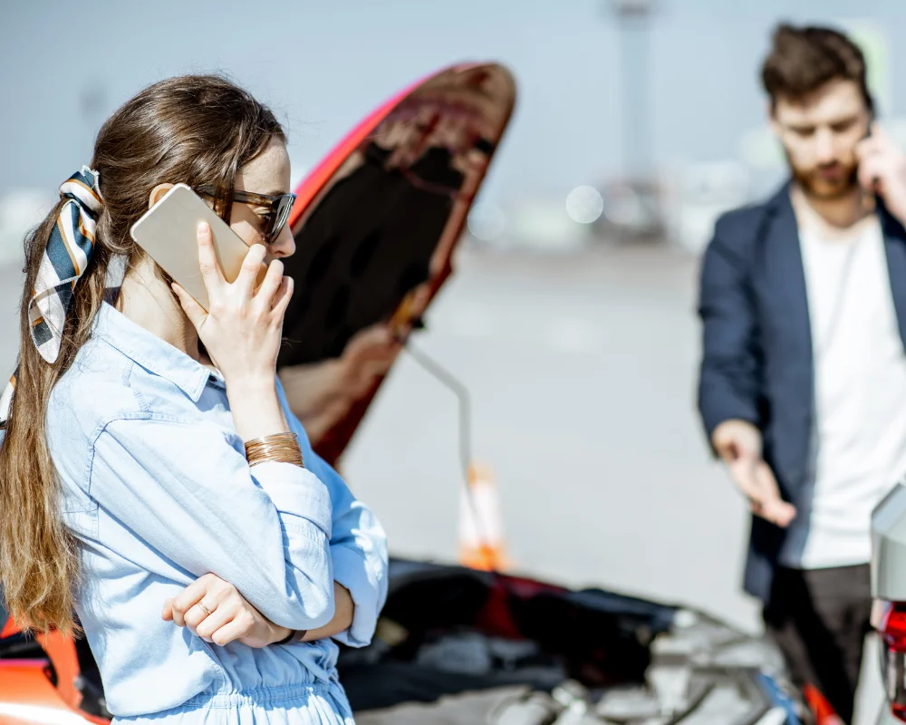 a woman in the foreground uses her cell phone while a man by two dented cars also uses his cell phone following a car accident