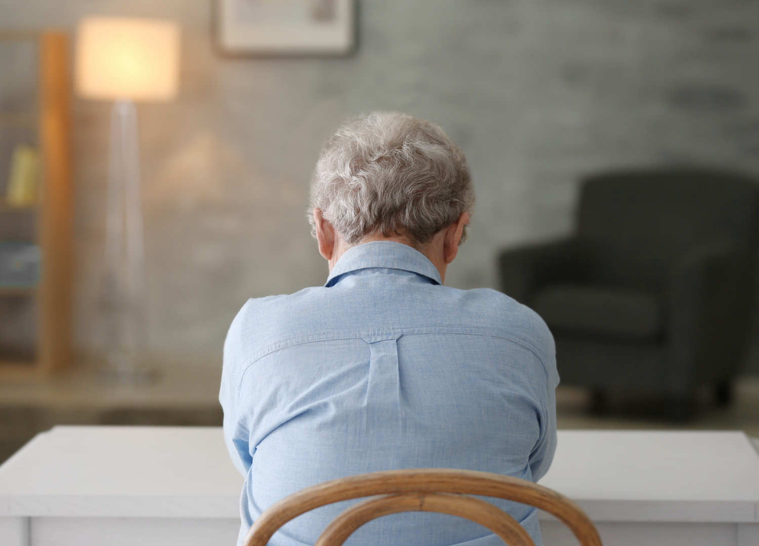 older woman sits at a table alone with her back to the POV of the camera, she appears to be sad judging by her body language, hunched shoulders and head down