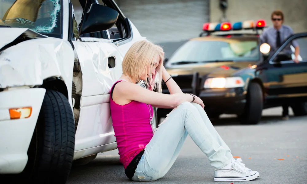 a young adult woman sits by her dented car, looking distressed, there is a cop car in the background, the woman may have a head injury