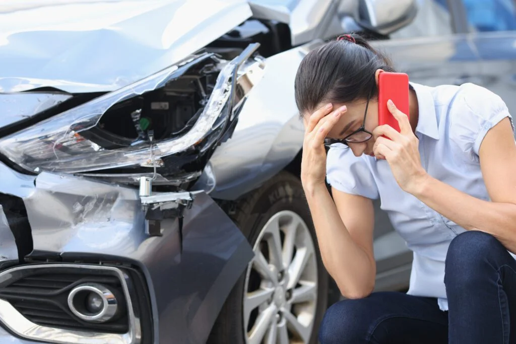 Una mujer de cabello oscuro usa el teléfono junto a un automóvil dañado recientemente en un accidente automovilístico.