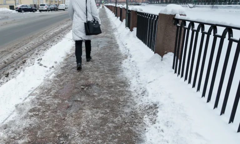 a woman in a winter coart faces away from the camera as she walks down a slippery sidewalk near a city street, snow surrounds the area