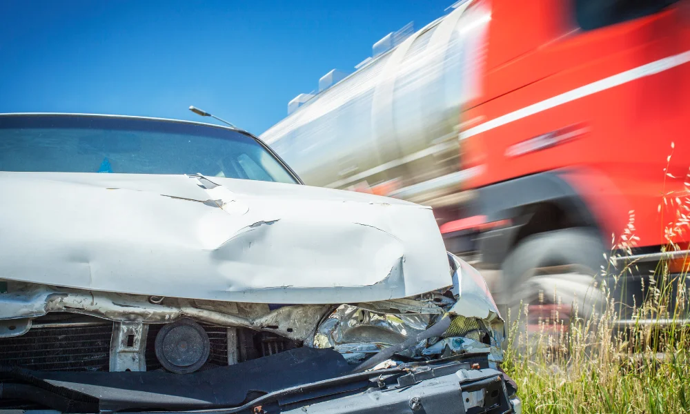 a white dented car in a grassy ditch after an accident with a semi-truck behind it