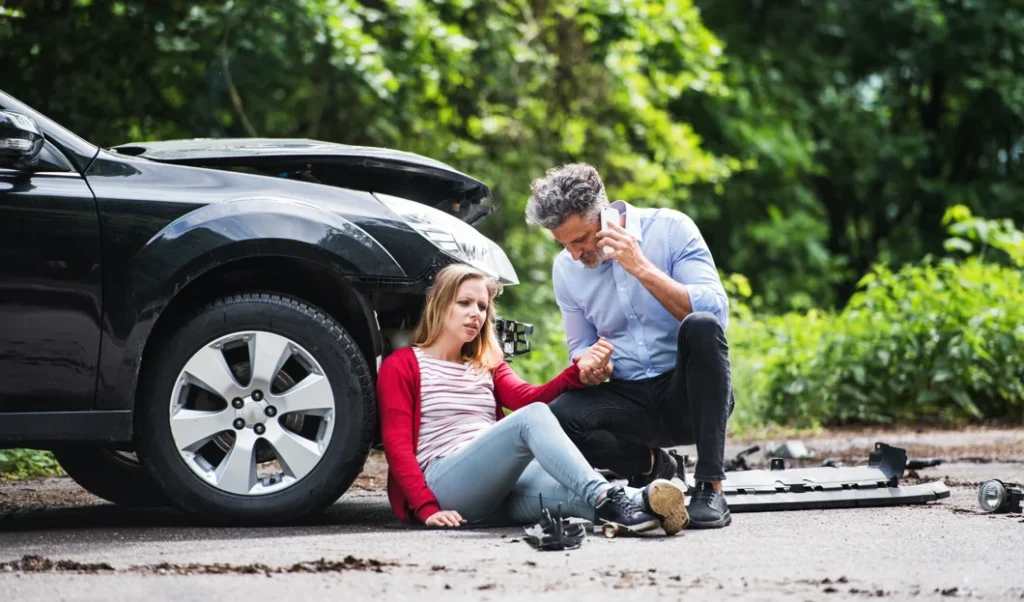 a woman appears injured on the side of the road, leaning against a damaged car, a man helps her