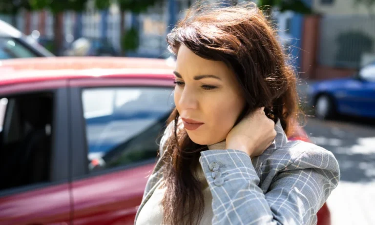 Una mujer está junto a un automóvil, sujetándose el cuello con dolor; parece confundida.