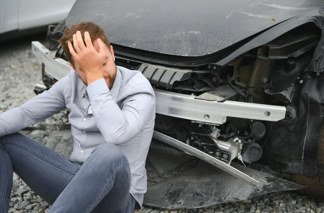 a confused man holds his head as he sits next to a damaged car post car accident