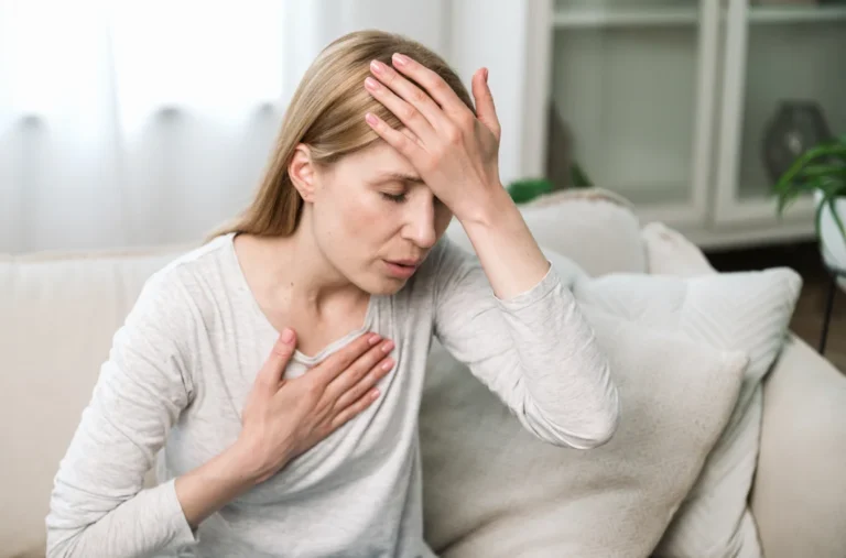 Una mujer rubia de mediana edad se sienta en su sofá con un dolor visible, sujetándose la cabeza y el pecho.
