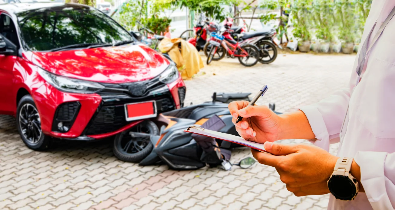 a person in the foreground fills out paperwork on a clip board, in the background is an accident scene with a car and a motorcycle